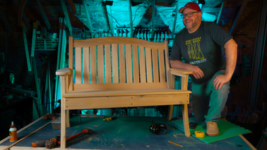 Matteout artisan builder smiling next to a handcrafted solid cedar garden bench on a workbench inside his woodworking shop.